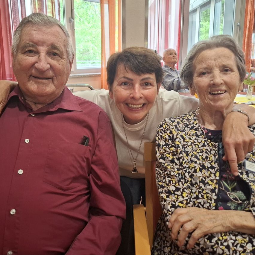 Three elderly individuals are smiling, two women and an older man, inside a room with windows and curtains.