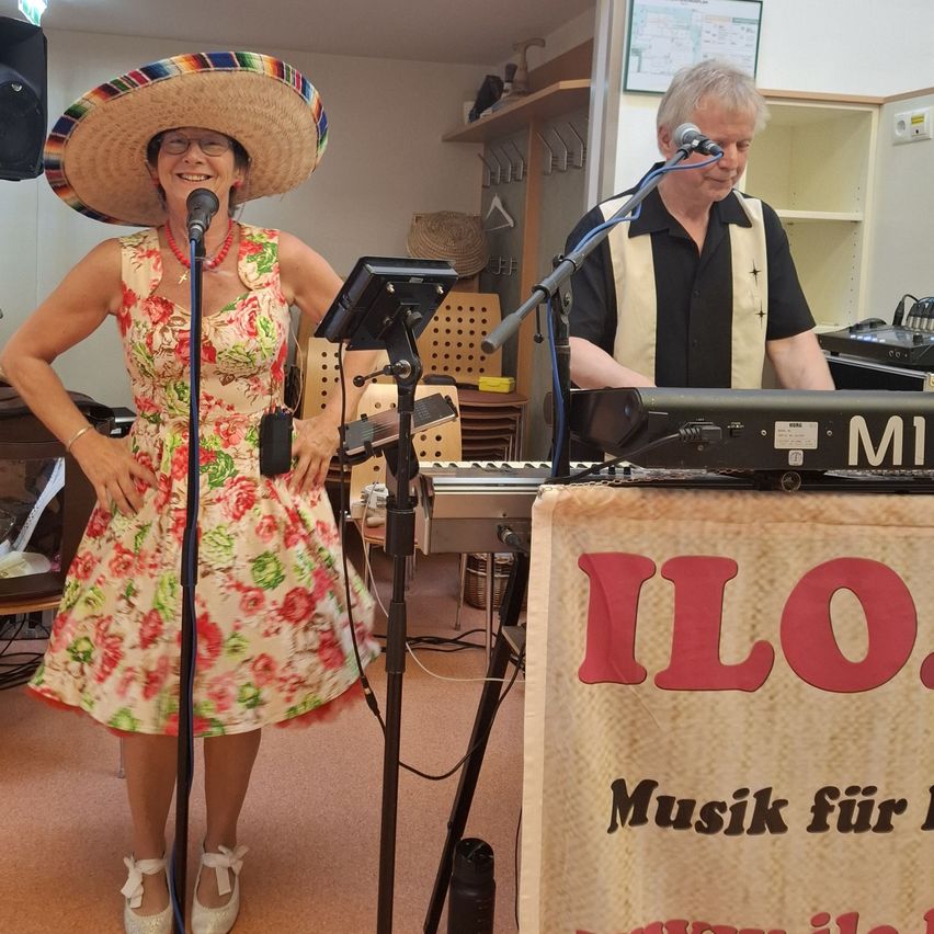 A woman in a floral dress and a sombrero stands next to a microphone. A man plays the keyboard behind her. The setting is a room with a banner that says 'ILO' and 'Music for you'.