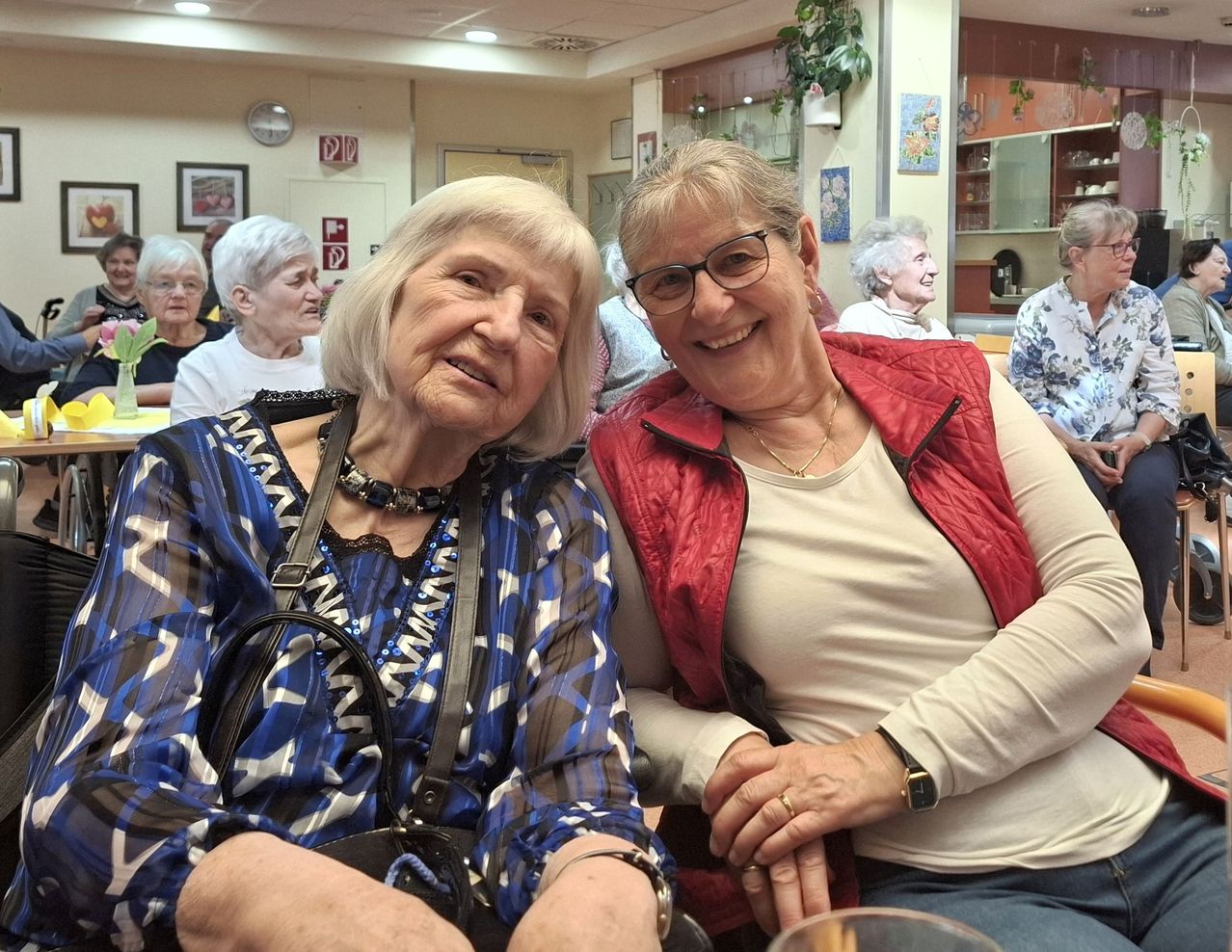 Two women, both elderly, are sitting next to each other in a room. They are smiling and appear to be posing for a photo.