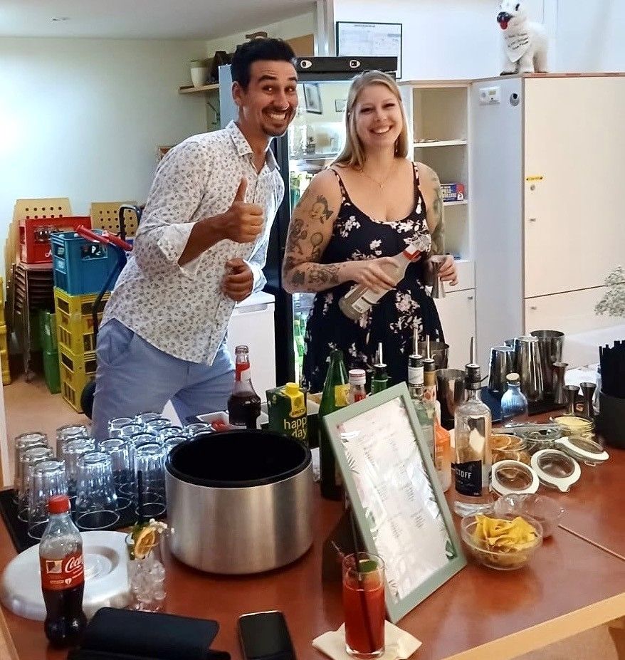 Ein Mann und eine Frau stehen hinter einer Bartheke mit verschiedenen Getränken und Flaschen und lächeln für ein Foto.