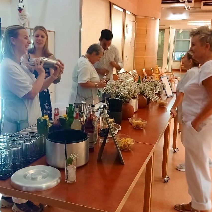 Eine Gruppe von Menschen in weißen Uniformen bereitet Getränke an einem Tisch mit verschiedenen Flaschen und Gläsern zu. Blumen und Snacks sind ebenfalls auf dem Tisch.