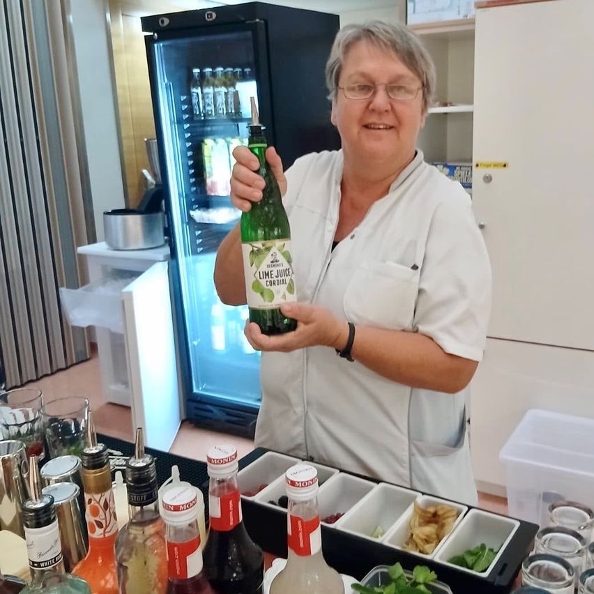 Eine Frau in einer weißen Laborkittel hält eine Flasche Lime Juice Cordial in einer Küche mit verschiedenen Gewürzen und einem Kühlschrank im Hintergrund.