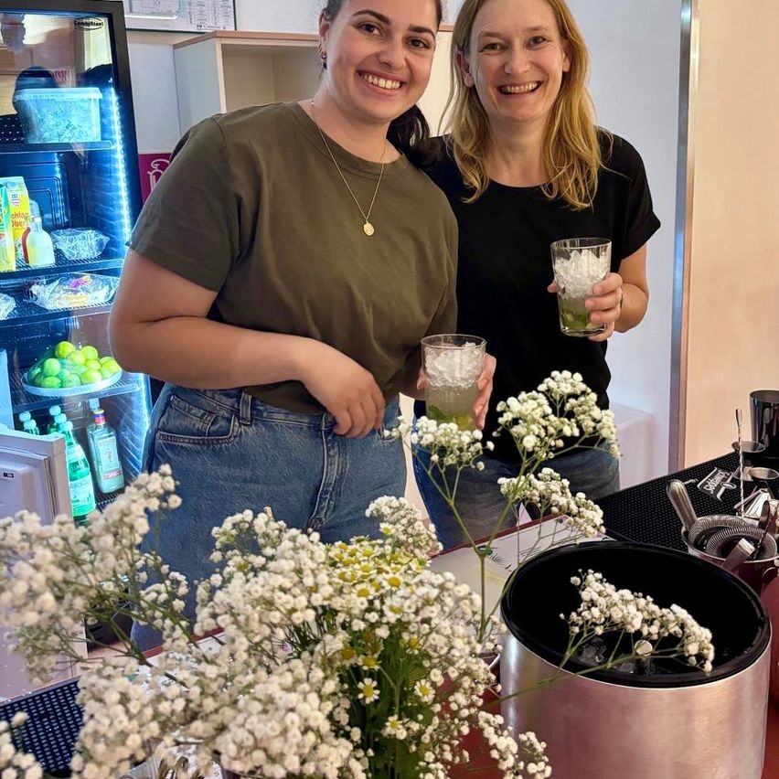 Zwei Frauen lächeln und posieren für ein Foto in einer Küche, eine hält ein Glas mit einem Getränk und die andere hält ein Glas mit Eis und einem Getränk. Es gibt Blumen auf dem Tisch und ein Kühlschrank mit Essen im Hintergrund.