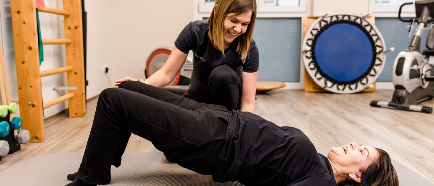 Eine Frau macht eine Rückenübung auf einer Matte, während eine andere Frau sie von hinten anleitet. Sie sind in einem Fitnessstudio.