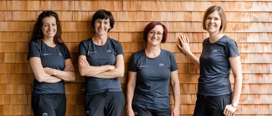 Vier Frauen in passenden Outfits stehen gegen eine Holzwand und lächeln für ein Foto. Jede trägt eine Brille und zwei haben ihre Arme verschränkt. Die Wand hinter ihnen besteht aus Holzschindeln.