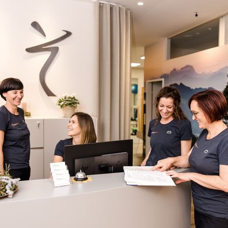 Vier Frauen, möglicherweise Mitarbeiterinnen, befinden sich in einem gut beleuchteten Empfangsbereich. Sie stehen vor einem Schreibtisch mit Monitor und Glocke. Die Frauen lächeln und scheinen mit einem Kunden zu sprechen. Die Wand hinter ihnen hat ein großes Bild von Bergen und eine dekorative Wandkunst.