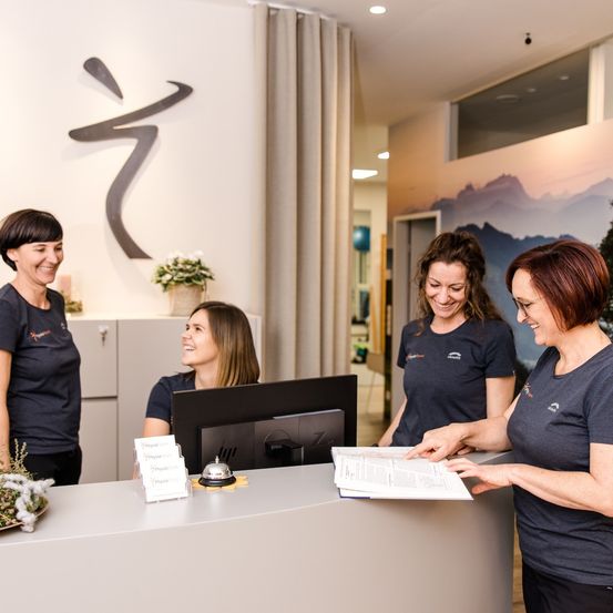 Vier Frauen, möglicherweise Mitarbeiterinnen, befinden sich in einem gut beleuchteten Empfangsbereich. Sie stehen vor einem Schreibtisch mit Monitor und Glocke. Die Frauen lächeln und scheinen mit einem Kunden zu sprechen. Die Wand hinter ihnen hat ein großes Bild von Bergen und eine dekorative Wandkunst.