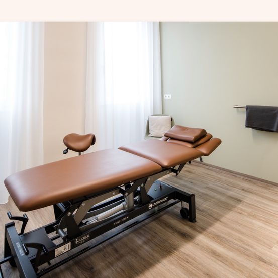 Ein leerer Raum mit Holzboden, einer hellen Wand und zwei braunen Massagetischen. Ein weißes Kissen und ein Handtuch befinden sich an der Wand.