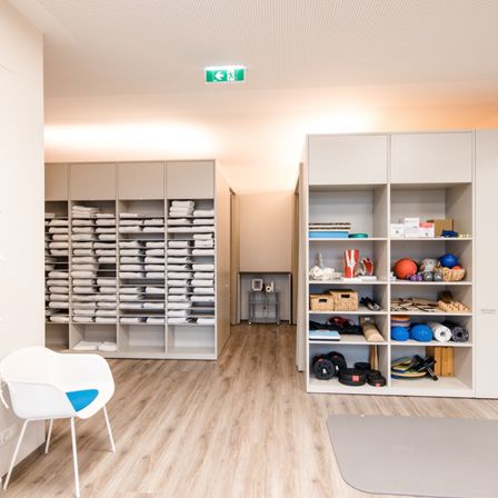 Ein Indoor-Fitnessstudio mit weißen Wänden, Holzböden und grauen Matten. Es gibt weiße Schränke, die mit gefalteten Handtüchern gefüllt sind, und ein weiterer Schrank mit verschiedenen Sportgeräten. Ein weißer Stuhl mit blauer Polsterung steht links. Ein Notausgangsschild befindet sich an der Decke.