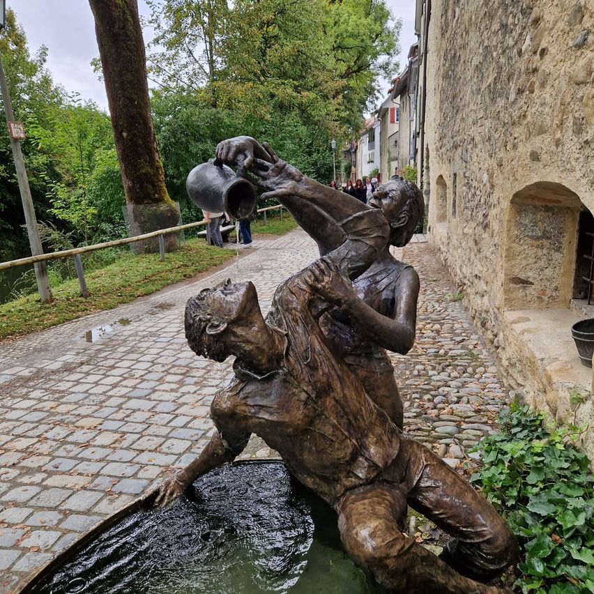 Eine Bronzeskulptur zeigt zwei Figuren in der Nähe einer Steinmauer und eines Wasserbrunnens auf einem Kopfsteinpflaster. Eine Figur gießt Wasser aus einem Krug auf die andere. Bäume und Gebäude sind im Hintergrund.
