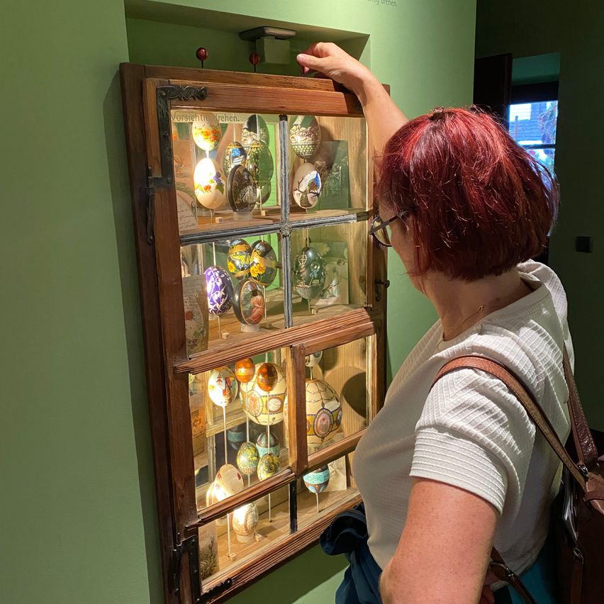 Eine Frau mit roten Haaren und Brille betrachtet eine Holzvitrine mit Glasfenstern, die dekorative Eier zeigt, während sie in einem Raum mit grünen Wänden steht.
