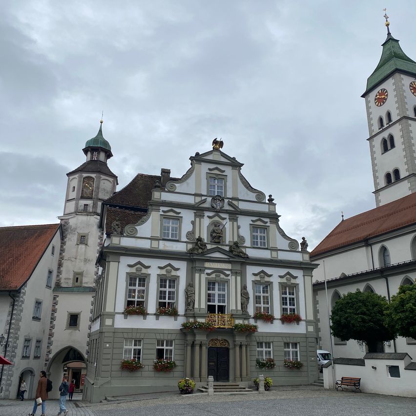 Ein großes, reich verziertes Gebäude mit einem Balkon und Blumenkästen an den Fenstern. Es hat zwei Türme, von denen einer eine Uhr hat. Eine Person geht unter einem roten Schirm.