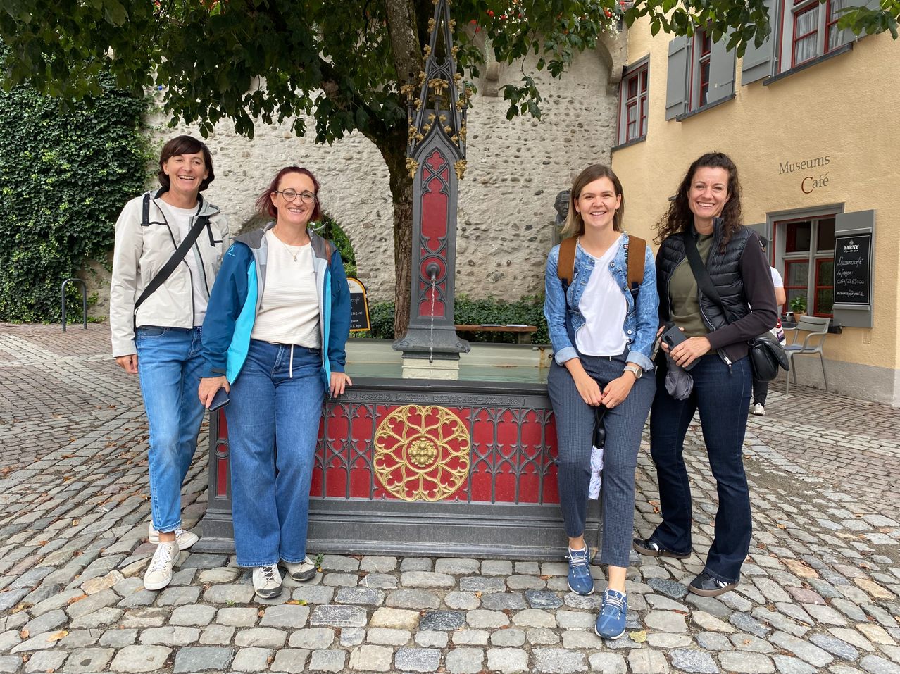 Vier Frauen lächeln und posieren für ein Foto vor einem Brunnen in einer historischen Stadt.