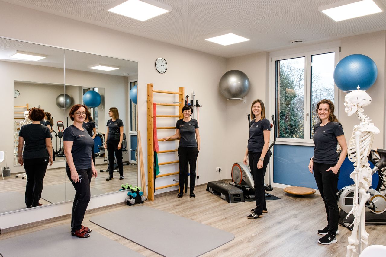 Fünf Frauen stehen in einem Fitnessstudio. Sie tragen schwarze T-Shirts und Hosen. Sie sind in der Nähe von Trainingsgeräten, darunter ein Spiegel, eine an der Wand montierte Uhr und ein großer Medizinball.