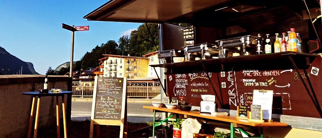 Ein Food Truck ist am Straßenrand geparkt mit einer Tafel mit Speisekarte und einem kleinen Tisch mit einer Flasche. Der Food Truck hat eine Tafel mit Speisekarte und Flaschen auf dem Regal.