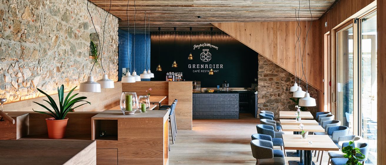 Das Interieur eines modernen Restaurants mit Holzboden, Wänden und Tischen, mit einer Steinmauer, einer Holzdecke mit hängenden Lampen und einer Bartheken mit dem Schild Grenadier Cafe Restaurant.