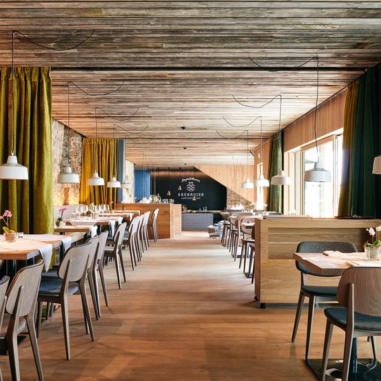 Das Innere eines Restaurants mit Holzböden, -wänden und -decke. Tische und Stühle sind in Reihen angeordnet, mit Lampen, die von der Decke hängen. Die Fenster haben Vorhänge und die Rückwand hat eine Tafel mit Schrift.
