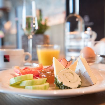 Ein Frühstücksteller mit Gurkenscheiben, Käse und Schinken steht auf einem Tisch mit einer Tasse, einem Weinkelch und einem Glas Orangensaft.