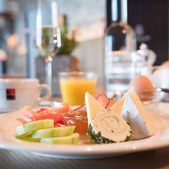 Ein Frühstücksteller mit Gurkenscheiben, Käse und Schinken steht auf einem Tisch mit einer Tasse, einem Weinkelch und einem Glas Orangensaft.