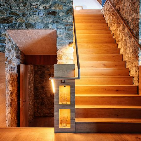 Eine Steinmauer mit einer Holztür führt zu einer Treppe aus Holz. Die Treppe hat ein Metallgeländer und zwei beleuchtete Nischen mit Gläsern.