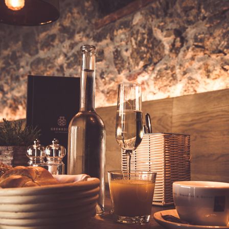 Ein Frühstückstisch mit einem Glas Orangensaft, einem Weinglas, einer Flasche und Brot auf einem Tisch mit einer Steinmauer im Hintergrund.