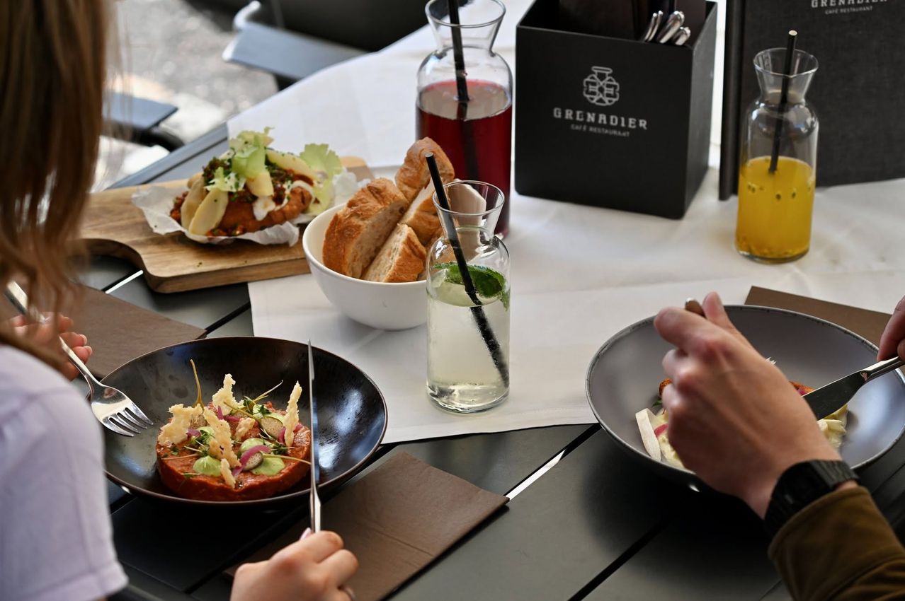 Two people sit at a table eating. Plates, bowls, and cutlery are on the table. A black box with the word 'Grenardier' is on the table.