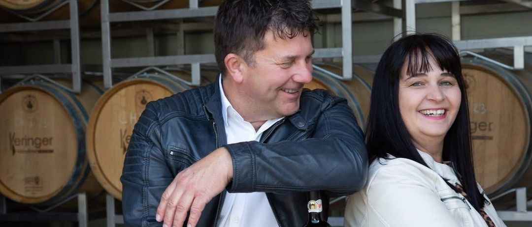 Ein Mann und eine Frau lächeln vor einer Reihe von Weinfässern mit der Aufschrift Leringer.