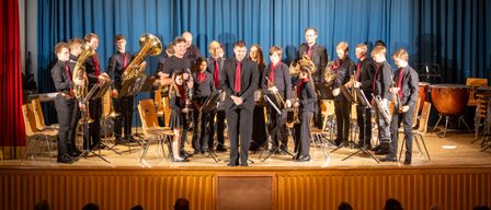 Ein Orchester spielt auf der Bühne mit verschiedenen Instrumenten, darunter Tubas und Trompeten, unter einem blauen Vorhang, während ein Publikum davor zuschaut.