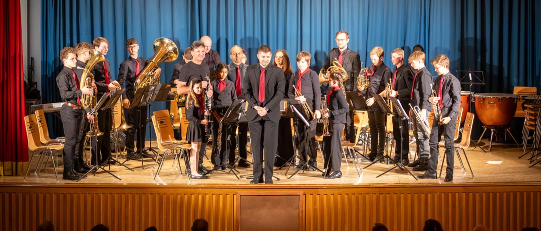 Ein Orchester spielt auf der Bühne mit verschiedenen Instrumenten, darunter Tubas und Trompeten, unter einem blauen Vorhang, während ein Publikum davor zuschaut.