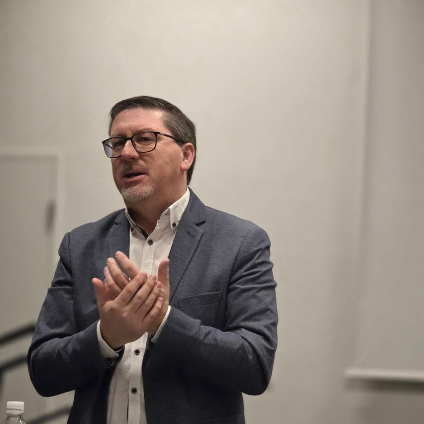 Ein Mann in einem grauen Anzug und mit Brille klatscht in der Hand, während er in einem Raum steht.