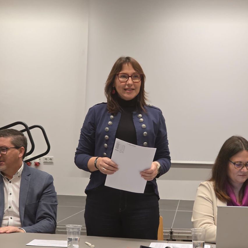 Drei Personen sitzen an einem Tisch in einem Konferenzraum. Eine Frau in der Mitte hält ein Blatt Papier. Der Mann auf der linken Seite trägt eine Brille und einen Anzug. Die Frau auf der rechten Seite trägt einen Schal und eine Brille.