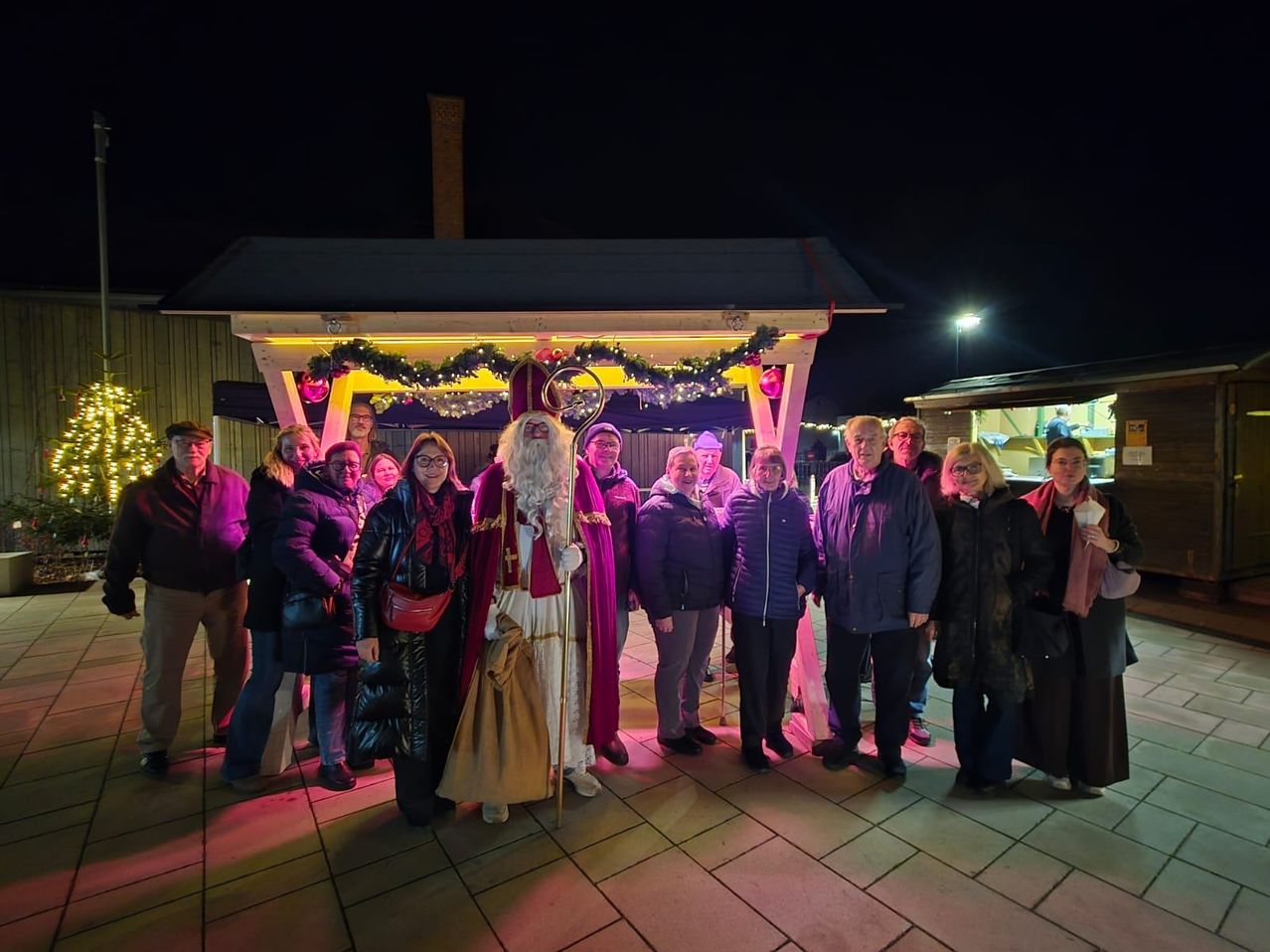 Eine Gruppe von Menschen in Winterkleidung steht bei Nacht vor einem geschmückten Pavillon, mit einer Person als Weihnachtsmann unter ihnen.
