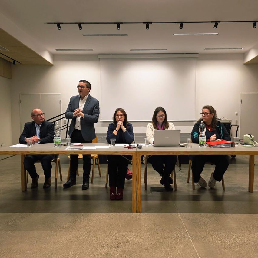 Eine Gruppe von Personen sitzt an einem langen Tisch in einem Raum. Ein Mann steht hinter dem Tisch und scheint zu sprechen.