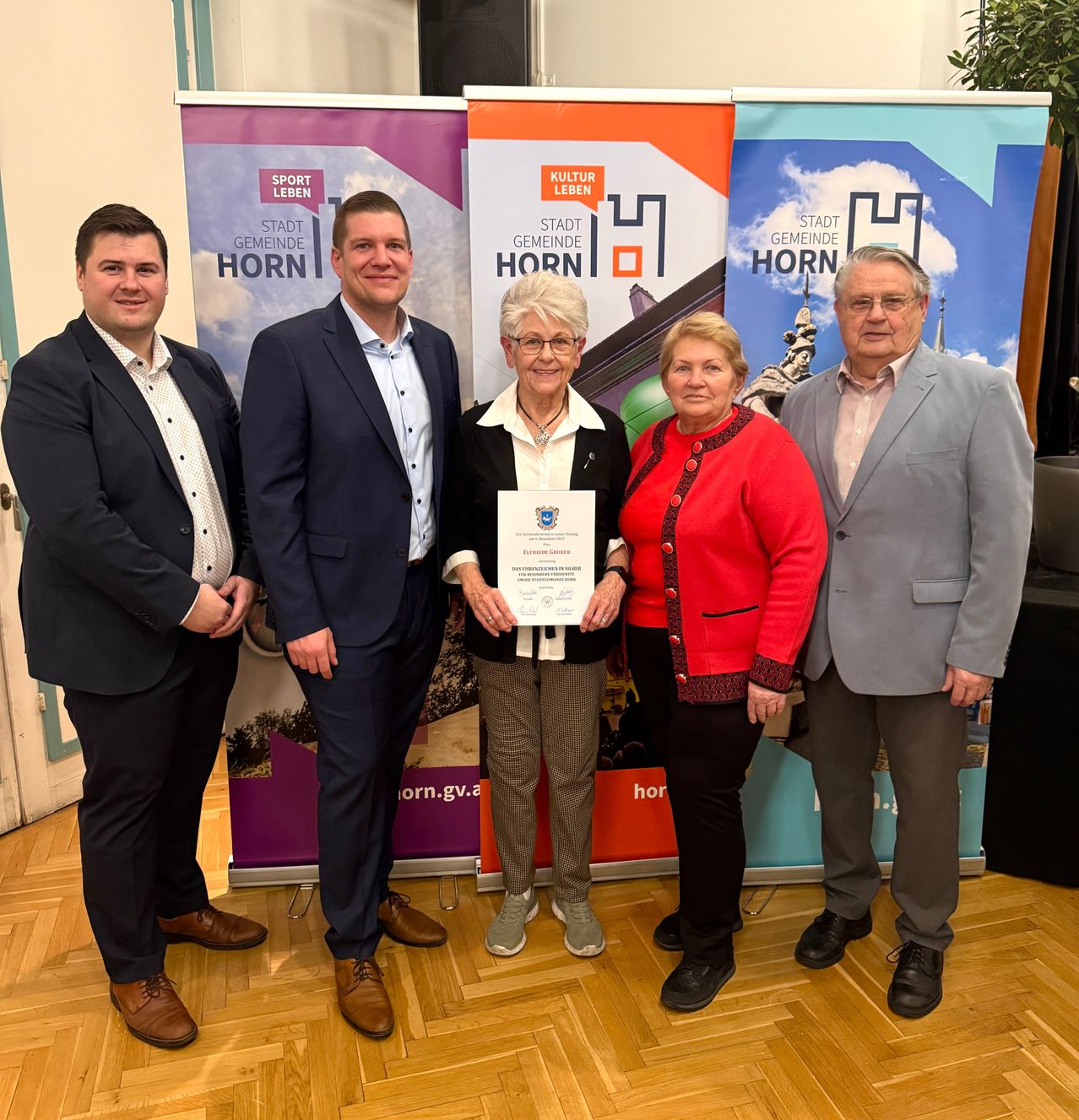 Fünf Personen stehen vor Bannern. Eine Frau in der Mitte hält ein Zertifikat. Hinter ihr stehen zwei Männer und zwei Frauen. Die Banner hinter ihnen haben die Worte 'Stadt Horn' darauf.