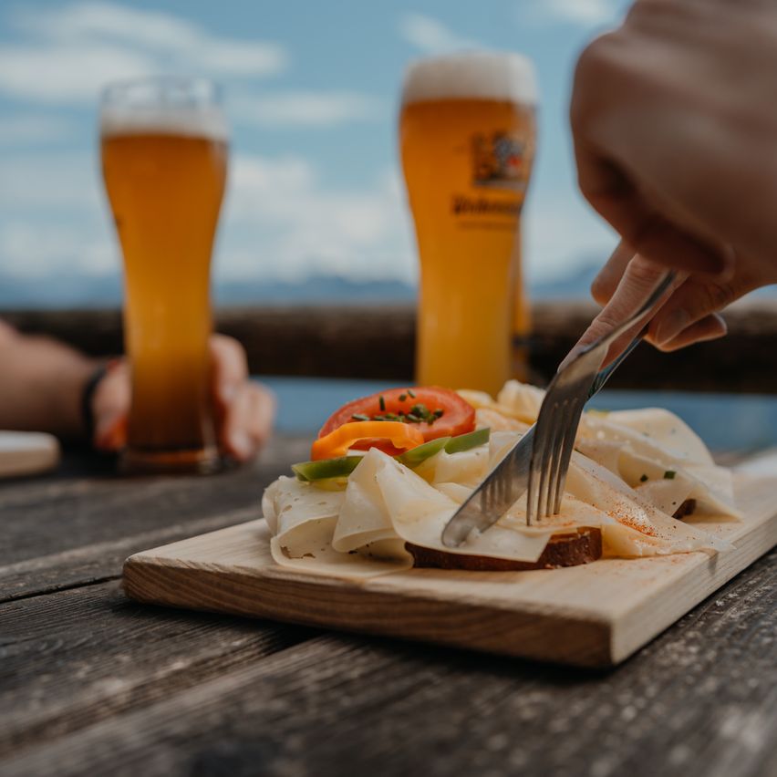 Eine Person schneidet ein Sandwich auf einem Holzbrett mit Messer und Gabel, mit zwei Gläsern Bier im Hintergrund.
