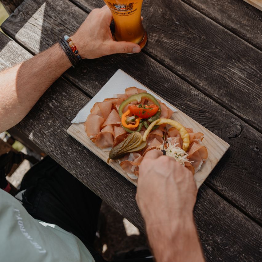 Eine Person hält ein Glas Bier und schneidet einen Teller mit Fleisch, Gemüse und Gurken auf einem Holztisch.