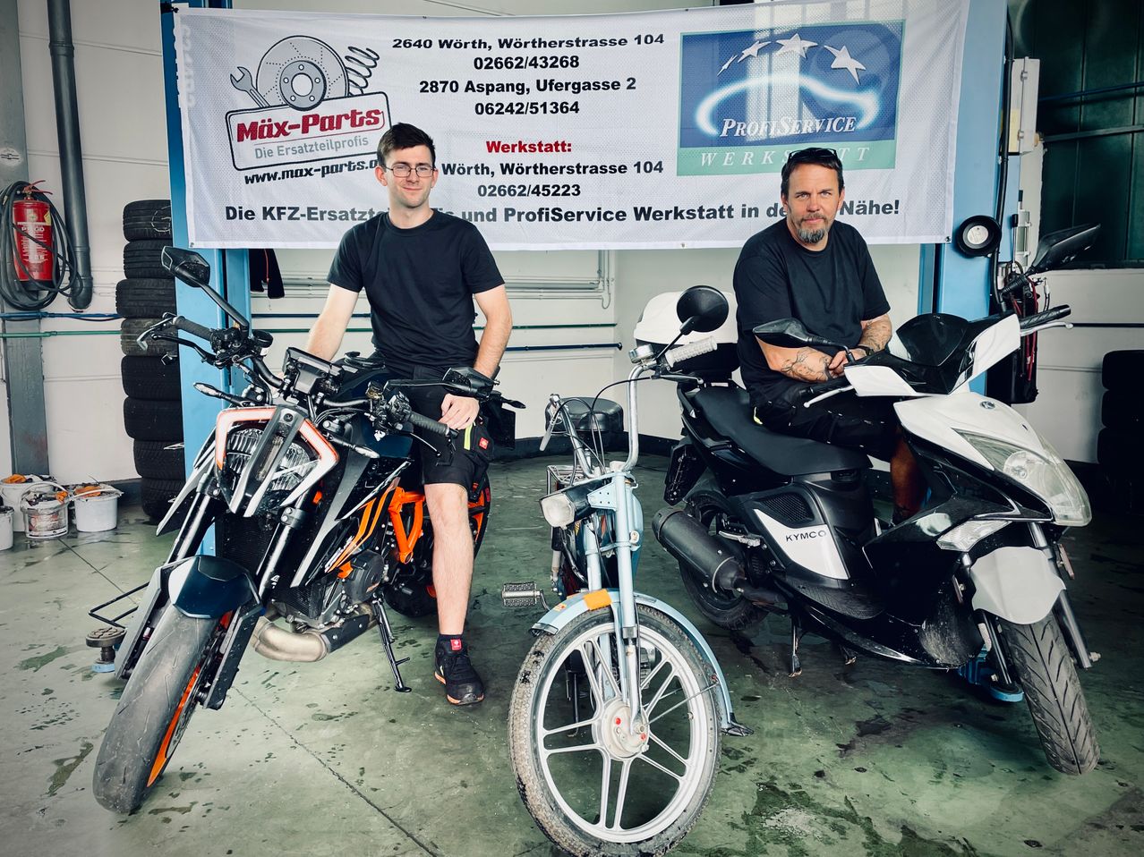 Zwei Männer sitzen auf Motorrädern in einer Werkstatt mit einem Banner, das Mix-Parts und ProfiService Werkstatt in der Nähe sagt. Ein Mann trägt eine Brille und Turnschuhe, der andere hat eine Tätowierung am Arm. Hinter ihnen ist eine Wand mit einem Poster.