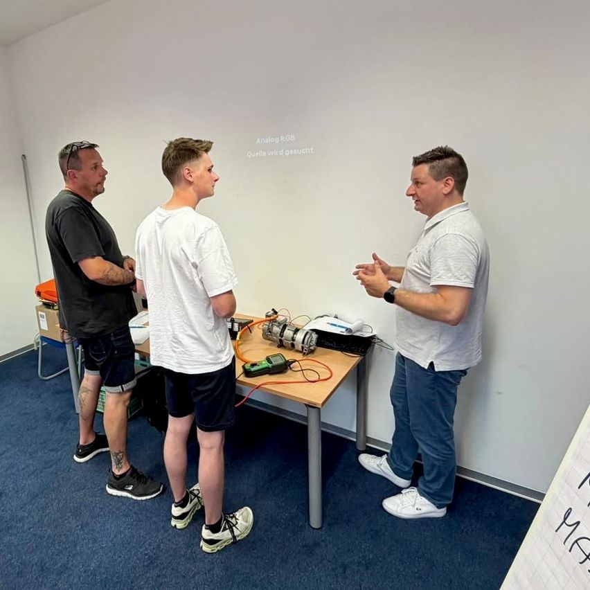Three men stand in a room, two wearing shorts and one with glasses on his head. They are near a table with a device, engaged in conversation. A whiteboard and text are on the wall behind them.