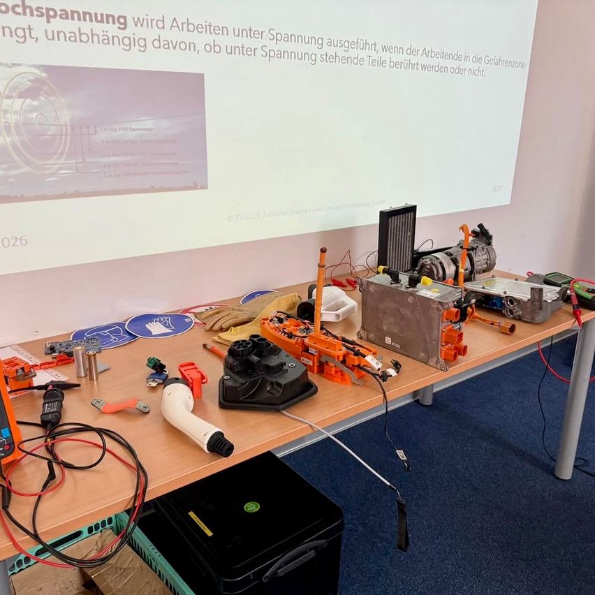 A table displays a variety of electronic equipment, including a power tool, a battery, and wires. Behind it, a projector screen shows a presentation about high voltage.