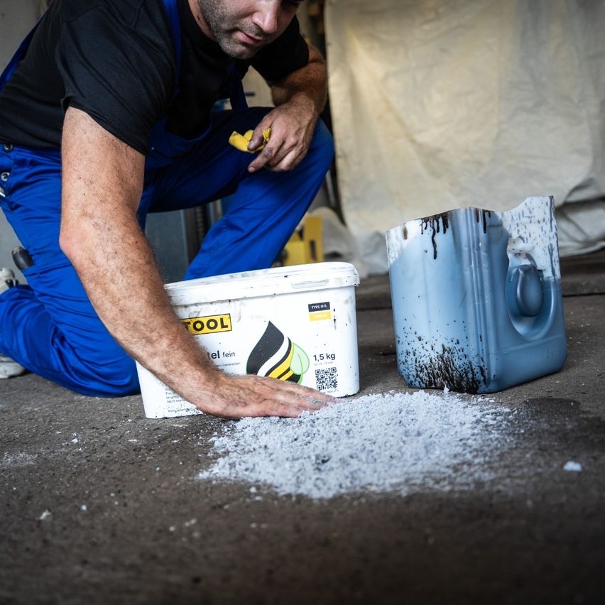 A man in work clothes is kneeling on the ground, holding a yellow cloth and a white bucket labeled 'TOOL'. He appears to be cleaning a spill. Nearby, a blue container with black stains is on the ground.