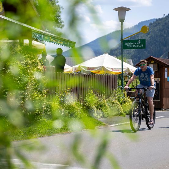 Ein Mann fährt mit dem Fahrrad die Straße hinunter vor einem kleinen Lokal mit einem grünen Vordach und einem Schild, das Steirisches Wirtshaus steht. Dahinter ist ein Gebäude mit einem Zeltdach zu sehen.