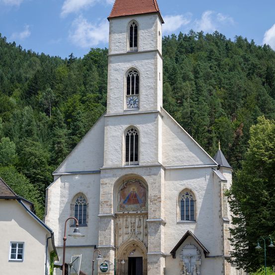 Eine weiße Kirche mit einem roten Dach und einer Uhr auf dem Turm steht vor einem mit Bäumen bedeckten Hügel.