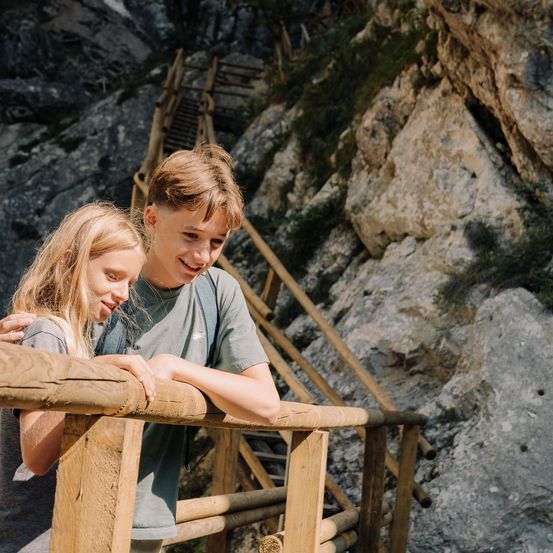 Zwei Kinder stehen auf einem hölzernen Geländer in einer Klippe mit einer Treppe an der Seite.