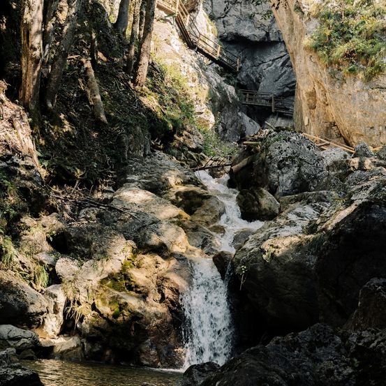 Ein Wasserfall in einem Gebirgsgebiet mit einer Holzbrücke und Felsen.
