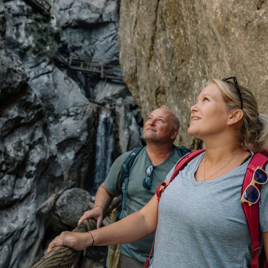 Ein Mann und eine Frau stehen auf einer Holzbrücke, vermutlich auf einem Berg. Die Frau schaut zum Himmel, der Mann schaut nach unten auf den Boden.