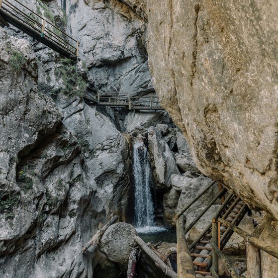 Ein Wasserfall, der einen felsigen Abhang hinunterströmt, mit Holzbrücken und Treppen, die darum herumführen.