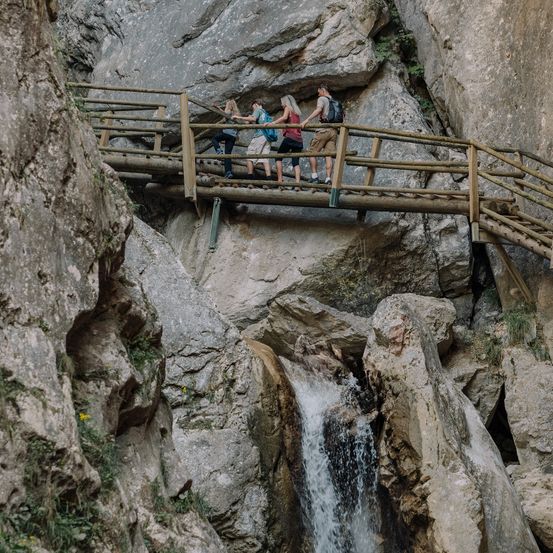 Vier Personen gehen auf einem Holzsteg über einen Wasserfall, drei von ihnen tragen Ruckscke und Turnschuhe.