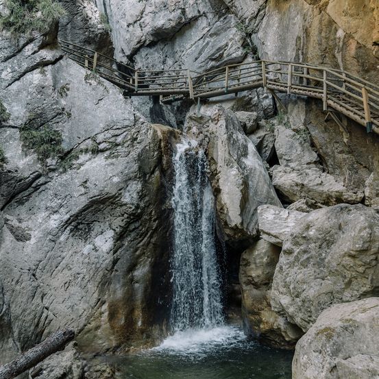 Eine Holzbrücke ist über einem felsigen Klippen errichtet, mit einem Wasserfall, der herunterfließt