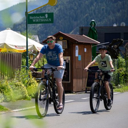 Ein Mann und ein Junge fahren auf einem asphaltierten Weg mit ihren Fahrrädern. Der Mann trägt eine Sonnenbrille und eine Uhr. Der Junge trägt eine Mütze und Turnschuhe. Hinter ihnen befindet sich ein Schild für ein Geschäft und ein Gebäude mit einer Speisekarte.