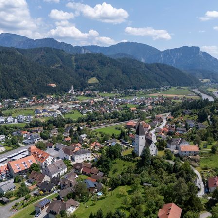 Eine Luftaufnahme eines Dorfes, umgeben von üppiger Vegetation und Bergen, mit einer Kirche und einem Fluss in der Ferne.